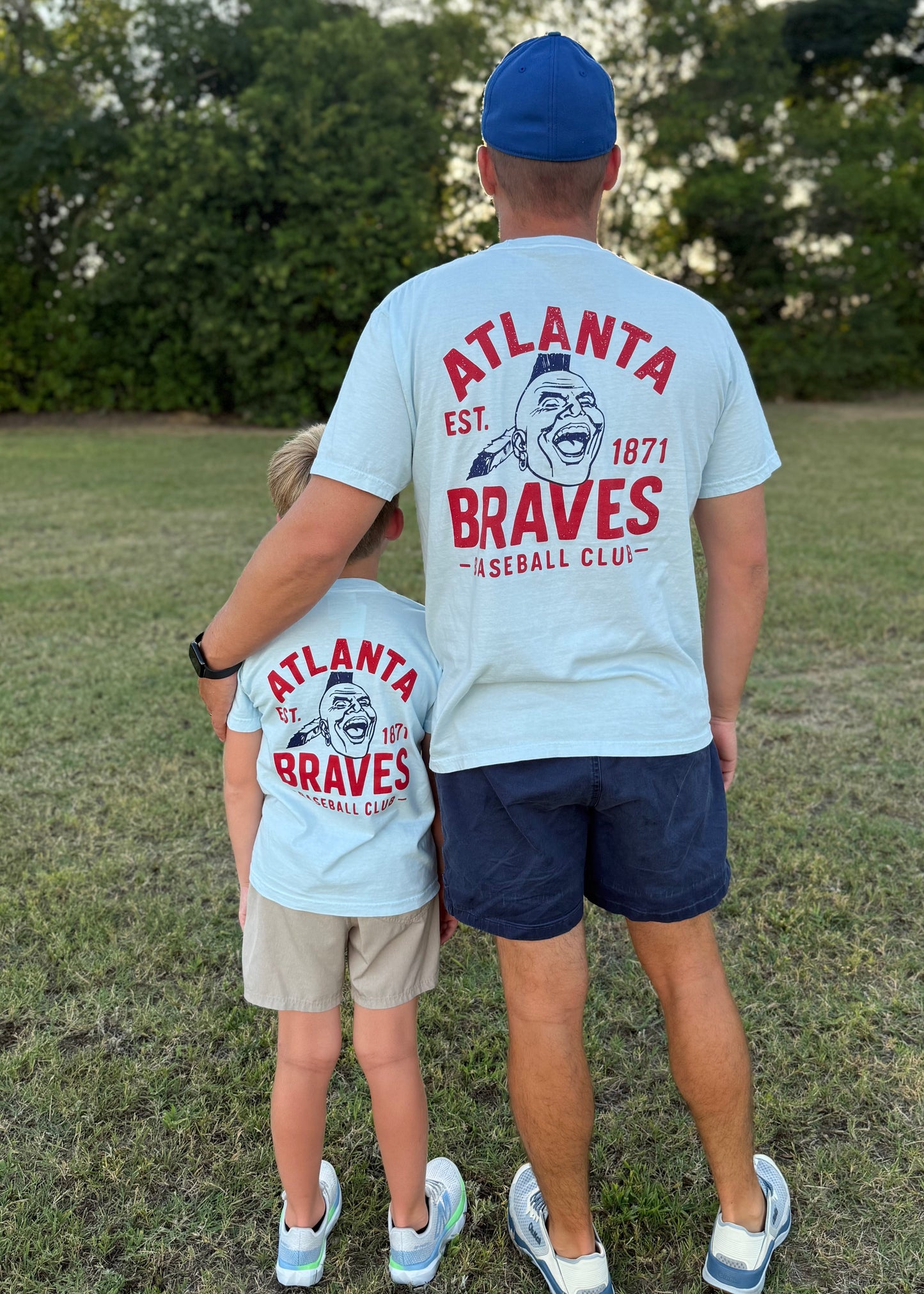 Two people wearing Atlanta Braves t-shirts standing on grass with trees in the background